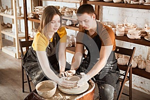 Couple in casual clothes and aprons making ceramic pot on pottery wheel in workshop.