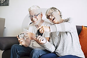 Couple of caucasian retired seniors playing videogames together at home sitting on the sofa - mature woman and man married - two