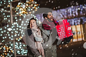 Couple in the city centre with holiday`s brights in background. Couple holding gift in a Christmas night