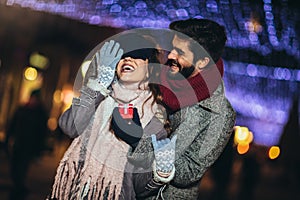 Couple in the city centre with holiday`s brights in background. Man presenting gift to woman