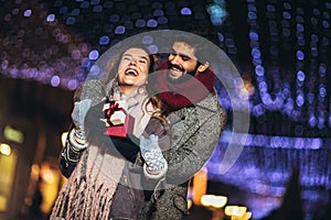 Couple in the city centre with holiday`s brights in background. Man presenting gift to woman