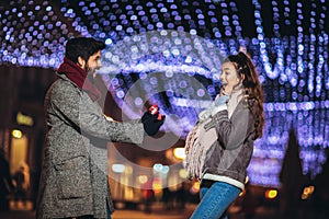 Couple in the city centre with holiday`s brights in background. Man presenting gift to woman