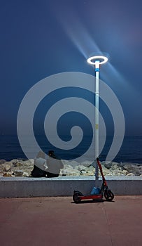 A couple on the city embankment under a street lamp and a parked electric scooter against the backdrop of the evening