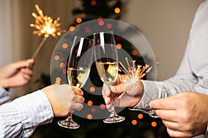 Couple clinking glasses with champagne and holding sparklers on background of christmas tree, celebrating christmas at home