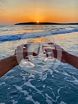 Couple clinking glasses of sea water on the beach at the background of scenic sunset over the sea. The sun goes below
