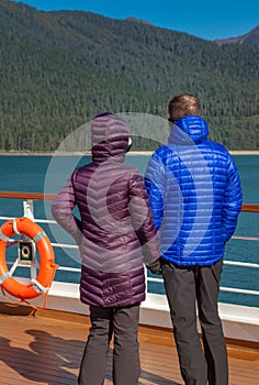 Couple in colorful purple and blue winter puffer jackets view Alaska, USA coastl from ship`s deck on a sunny cold day.