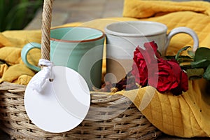 Couple cups of coffee in a basket with blank tag paper label on yellow background. Mugs ceramic with two red roses, rattan basket.