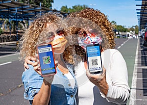 Couple of curly and blonde women ready to travel showing digital health certification for people vaccinated of coronavirus