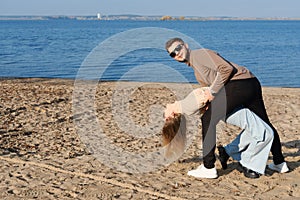 The couple is dancing, having fun and fooling around on the beach. A young bearded man in sunglasses hugs a girl by the