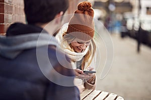 Couple On Date Sitting Outside Coffee Shop On High Street Using Mobile Phone