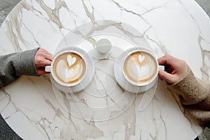 Couple on a date, two people, hands holding coffee cup with latte art. Cappuccino crema of heart shape. Top view of marble cafe