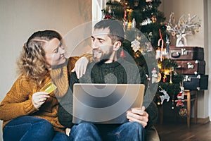Couple doing Christmas shoppings online on the sofa at home with laptop, looking at each other and smiling