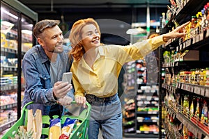 Couple doing grocery shopping together, man using cellphone, woman pointing at shelf, choosing food products in