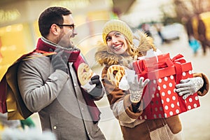 Couple dressed in winter clothing holding gift boxes outdoor. They eat pretzels