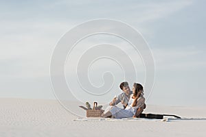 Couple drinking champagne while sitting on blanket with basket of fruits and acoustic guitar on beach