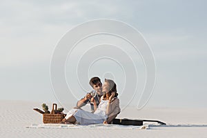 Couple drinking champagne while sitting on blanket with basket of fruits and acoustic guitar on beach