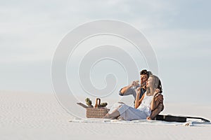 Couple drinking champagne while sitting on blanket with basket of fruits and acoustic guitar on beach