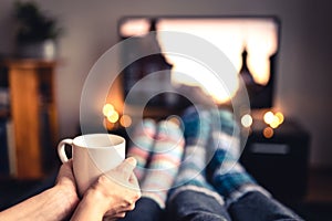 Couple drinking tea, hot chocolate, eggnog or mulled wine and watching tv in warm cozy woolen socks in winter.