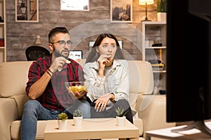 Couple eating chips and looking shocked at tv while watching a movie