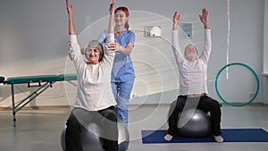 couple of elderly patients doing exercises while sitting on fitballs, nurse helping grandparents raise their arms during
