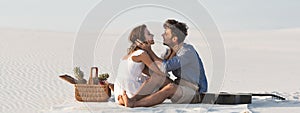 Couple embracing while sitting on blanket with basket of fruits and acoustic guitar on beach