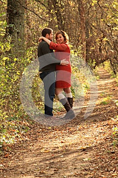 Couple embracing on trail in fall wooded area as woman coyly smiles at camera
