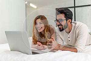 Couple enjoy leisure time at home using laptop computer laying on bed surfing internet together.