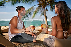 Couple enjoys fresh coconut water on tropical beach. Man, woman sip cocktails, relax under palm trees by sea. Romantic