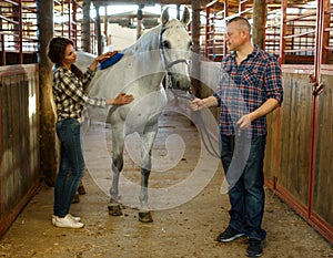 Couple of farmers cleaning horse while standing at stabling indoor