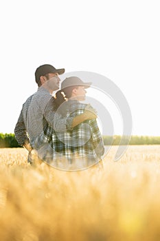 A couple of farmers in plaid shirts and caps stand embracing on agricultural field of wheat at sunset.