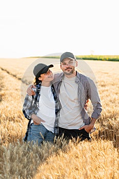 A couple of farmers in plaid shirts and caps stand embracing on agricultural field of wheat at sunset.