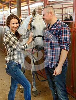 Couple of farmers standing with white horse at stabling indoor