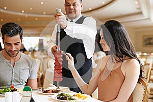 Couple, fine dining and waiter serving food or sausage kebab on skewer or restaurant, anniversary or nutrition. Man