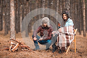 A couple in the forest. A girl holds a mobile , a guy sharpens a wooden bar with a knife.
