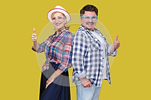 Couple of friends, adult man and woman in casual checkered shirt standing together back to back, showing thumbs up, toothy smile