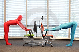 Couple in full body elastic suits exercising with chairs in office