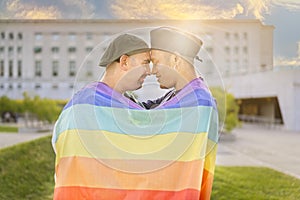 Couple of gay men in love embracing each other, wrapped in a gay flag, in a park at sunset, with reflections of the sun.