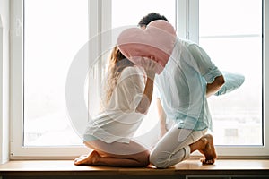 Couple girl and guy play with pillows near window.