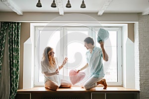 Couple girl and guy play with pillows near window.