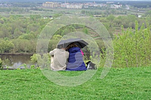 A couple of guy and girl are sitting on the green grass on the bank of the Klyazma river under one umbrella and are