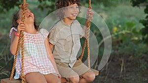 Couple of happy kids hugging and swinging on wooden swing at golden sunset. Brother embracing little sister on a swing