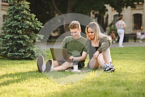 Couple of happy students using laptop while sitting on university campus lawn, looking at screen and smiling. Smiling girl and