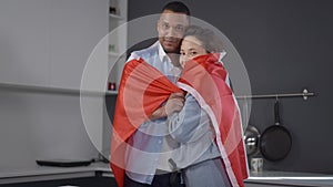 Couple of happy young Canadians wrapping in national flag looking at camera smiling. Portrait of loving African American