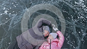 Couple has fun during winter walk against background of ice of frozen lake. Lovers lie on clear ice, have fun, kiss and