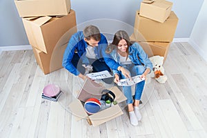 Couple have a discussion about design of their new home sitting on the wooden floor between many cardboard boxes