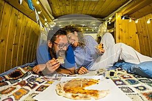 Couple have fun eating pizza inside vintage hand made wooden van vehicle in alternative lifestyle and travel vacation - happy