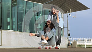 Couple have fun on longboard: male push back of joyful female sitting on skateboard and laughing