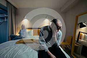 Couple have problems in bedroom. Frustrated man sits on edge of bed, angry woman behind. Impotence and other male health