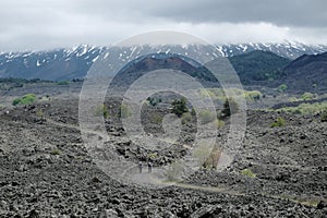 Couple hikers on the path to extinct cinder cones under the majestic volcanic cone of Etna Mount that is covered by clouds