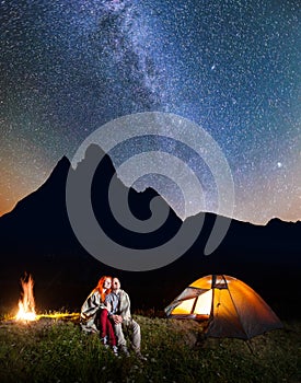 Couple hikers sitting together near campfire and glowing tent at night under stars and looking to the starry sky